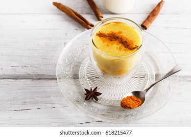 Close up image of golden latte in a transparent glass, spices and bottle of milk over white wooden table 