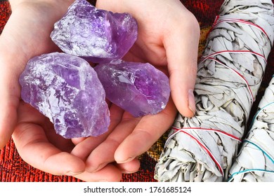 A close up image of a hand holding three purple amethyst crystals next to two white sage bundles. 