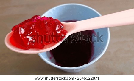 Close up image of an individual serving of red fruit flavored jelly, Strawberry jelly-cup, cherry-jelly, raspberry gel, in a plastic container with food in the plastic spoon. Fun,  Individual mixed red fruits jelly cup, single-serve jelly, snack cup, colorful kid's favorite dessert treat a strawberry, raspberry, or red fruit-jelly. Single-portion, Convenient, Ready-to-eat, old fashioned, made without gelatine kid's favorite dessert, in a plastic serving dish. Photograph of plant-based gelling agents, Vegan-jelly, made with agar-agar or pectin. Individual serving of a red vegan jelly in a cup. Jello or jello cup.