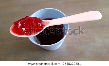 Close up image of an individual serving of red fruit flavored jelly, Strawberry jelly-cup, cherry-jelly, raspberry gel, in a plastic container with food in the plastic spoon. Fun,  Individual mixed red fruits jelly cup, single-serve jelly, snack cup, colorful kid's favorite dessert treat a strawberry, raspberry, or red fruit-jelly. Single-portion, Convenient, Ready-to-eat, old fashioned, made without gelatine kid's favorite dessert, in a plastic serving dish. Photograph of plant-based gelling agents, Vegan-jelly, made with agar-agar or pectin. Individual serving of a red vegan jelly in a cup. Jello or jello cup.