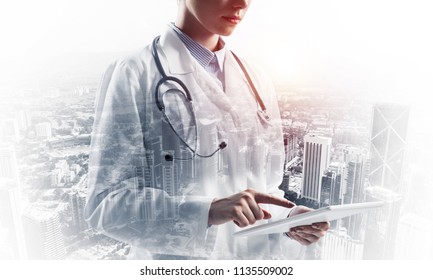 Close up image of medical employee in white suit using tablet and standing outdoors with city view on background. Medical industry concept. Double exposure