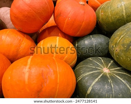 A close up image of a pile of pumpkins. The pumpkins are mostly orange, with a few green ones mixed in. The pumpkins are all different sizes and shapes.