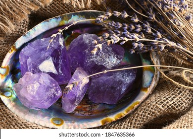 A close up image of several rough pieces of amethyst crystal in an abalone shell on vintage burlap fabric. 