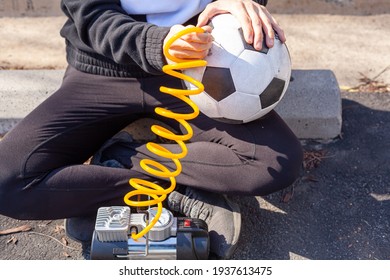 Close up image showing a caucasian woman holding a soccer ball and inserting needle bit at the end of the yellow coiled tubing attached to 12v car tire inflator to pump in air to the ball. 