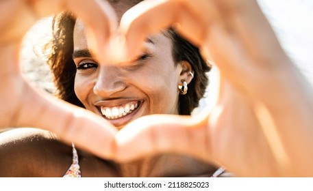 Close up image of smiling woman in swimwear on the beach making a heart shape with hands - Pretty joyful hispanic woman laughing at camera outside - Healthy lifestyle, self love and body care concept - Shutterstock ID 2118825023
