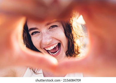 Close up image of smiling woman in swimwear on the beach making a heart shape with hands - Pretty joyful woman laughing at camera outside - Healthy lifestyle, self love and body care concept