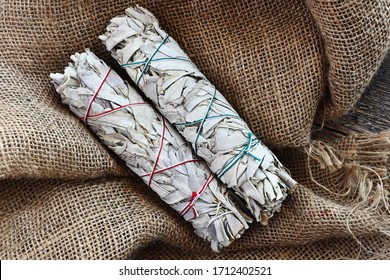 A close up image of two white sage smudge sticks on beige burlap fabric.  