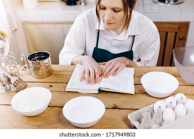 Close Up Image Of Woman Sitting At Wooden Kitchen Table Looking To  Recipe Book And Trying To Choose What To Cook. Cooking At Home Concept, Lifestyle. Ketogenic Diet And Menu