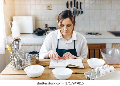 Close Up Image Of Woman Sitting At Wooden Kitchen Table Looking To  Recipe Book And Trying To Choose What To Cook. Cooking At Home Concept, Lifestyle. Ketogenic Diet And Menu