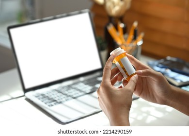 Close Up Image Of A Woman's Hand Holding A Pill Bottle To Read The Drug Label With The Laptop With Empty Screen As The Background.