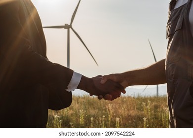 Close up of indian technician and african american inspector shaking hands while standing on farm with wind turbines. Concept of people, teamwork and eco friendly energy.