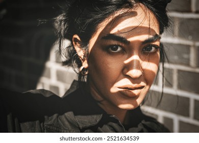 Close up of Indian woman with dark hair and hoop earrings. The Indian woman has a serious expression. Shadows play across woman's face. Indian woman is standing against brick wall close up portrait.