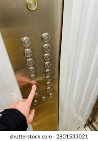 Close up inside elevator shows hand pressing fifth floor button on steel control panel. Polished chrome circles reflect light against rugged metal surface, white wall panels framing compact space