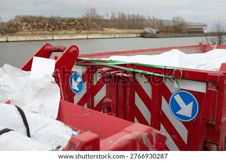Close interior view of red skip container with covered materials secured by strap. Industrial port environment with canal in background.