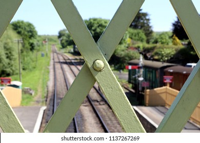 Close up of the ironwork of an old railway footbridge, with track, platform, signals and rolling stock in the blurred background, on a sunny summer day