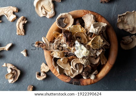 Close up isolated flat lay image of a rustic wooden bowl filled with sliced and dried mushroom pieces. The bowl is on dark stone surface. There is an assortment of mushroom types in this moody image.
