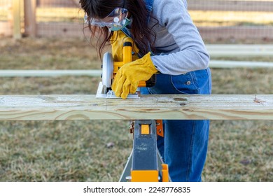 Close up isolated image of a construction worker or carpenter operating an electrical circular saw to slice a thick pressure treated wooden plank on a sawhorse. She wears overalls, safety googles