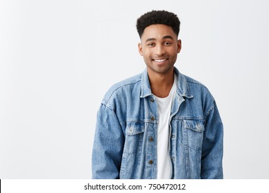 Close up isolated on white portrait of young beautiful cheerful dark-skinned male university student with afro hairstyle in white t-shirt and denim jacket smiling brightly in camera. Copy space