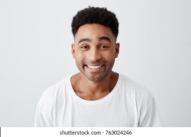 Close up isolated portrait of cheerful happy young man with afro hairstyle in casual white t-shirt smiling brightly, looking in camera with excited and joyful expression.
