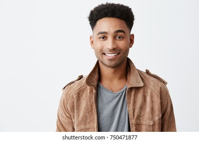 Nahaufnahme, isoliertes Porträt eines jungen, dunkelhäutigen und attraktiven Mannes mit afro-Haarstil in grauem T-Shirt unter brauner Jacke, der mit Zähnen lächelt, die in der Kamera mit fröhlichem und friedlichem Gesichtsausdruck aussehen.