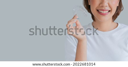 close up (isolated) young beautiful asian woman smiling with hand holding dental aligner retainer (invisible) on gray background of dental clinic for beautiful teeth treatment course concept