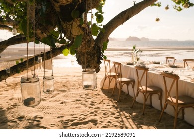 Close Up The Jar Candle Hanging On The Tree With Long Table Wedding Dinner Setup On The Beach At Thailand In The Evening. Wedding Party Concept. Decoration Outdoor Restaurant At The Beach. 