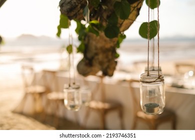 Close Up The Jar Candle Hanging On The Tree With Long Table Wedding Dinner Setup On The Beach At Thailand In The Evening. Wedding Party Concept. Decoration Outdoor Restaurant At The Beach.