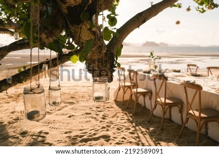 Close up the jar candle hanging on the tree with long table wedding dinner setup on the beach at Thailand in the evening. Wedding party concept. Decoration outdoor restaurant at the beach. 