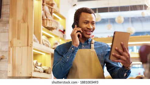 Close up of joyful African American young handsome man worker in apron stands in bakery shop, speking on smartphone and using tablet device. Business concept. Retail industry