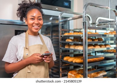 Close up of joyful African American young woman worker in apron stands in bakery shop, speking on smartphone and using tablet device. Small business concept. - Shutterstock ID 2145805139
