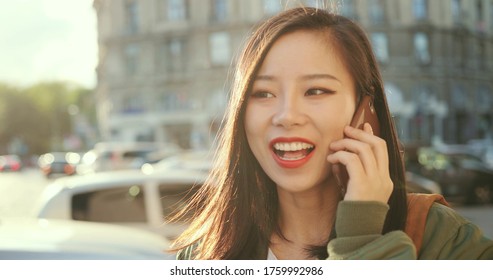 Close Up Of The Joyful Asian Young And Pretty Girl Talking At The Phone And Laughing At The Noisy Street Of The Town.