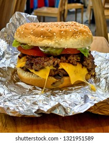 Close Up Of Juicy Cheeseburger In Burger Joint, Unwrapped Hamburger On Foil On Restaurant Table In USA, American Fast Food, Patty With Melting Cheese And Salad