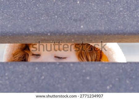 Close up of kid looking through gap in wooden fence. Curious red haired child peeking with eyes wide open. Playful childhood moment outdoors at home. Concept of discovery and exploration.