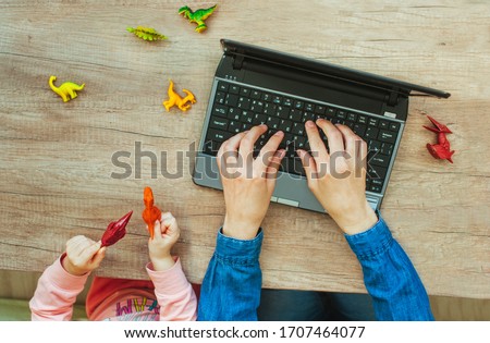 Close up of kids hands playing with dinosaur toys while sitting near mother typing on the laptop on wooden background. Work from home during quarantine concept. Top view, flat lay.