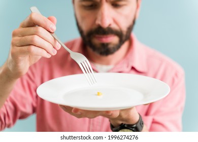 Close Up Of A Latin Man Holding A Plate And A Fork With Very Little Food. Depressed Man Can't Eat Due To His Bad Mental Health And Eating Disorder