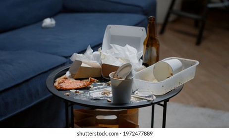 Close Up Of Leftover Food On Table In Empty Messy Living Room, Bottle Of Beer And Napkins On Blue Sofa. Unorganized Apartment Of Alone Woman With Sever Depresion Having Trash Rubbish With No People In