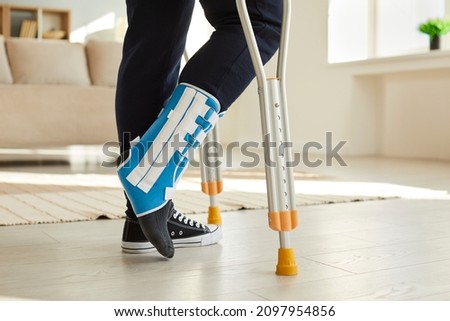 Close up leg of man walking with crutches in ankle brace with support adjustable strap fracture fixator. Cropped image of low angle leg of man who has leg injury and walks with crutches room at home