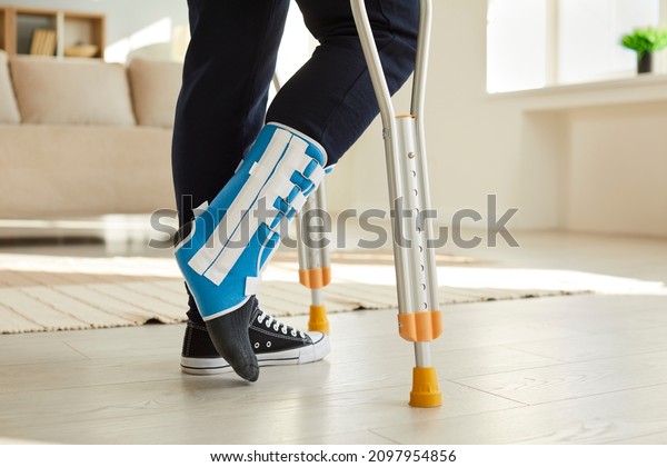 Close up leg of man walking with crutches in ankle
brace with support adjustable strap fracture fixator. Cropped image
of low angle leg of man who has leg injury and walks with crutches
room at home