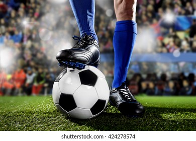 Close Up Legs And Feet Of Football Player In Blue Socks And Black Shoes Standing  With The Ball Playing Match At Soccer Stadium On Green Grass Pitch With Audience Flash Lights And Flare