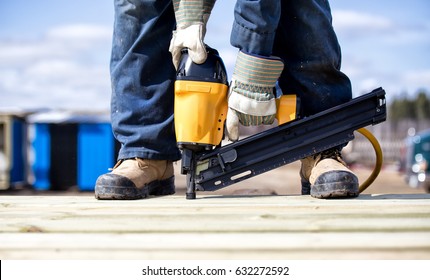 Close Up Of Legs And Feet In Steel Toe Boots And Arms Of Man In Overalls And Work Gloves Using An Air Nailer On Wood Boards Outdoors In Summer