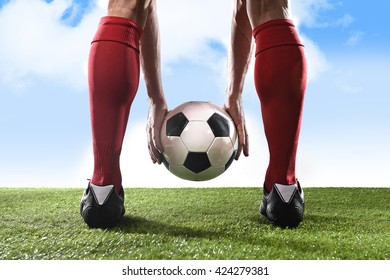 close up legs of football player in red socks and black shoes holding the ball in his hands placing it at the free kick or penalty spot playing outdoors on green grass pitch under a blue sky