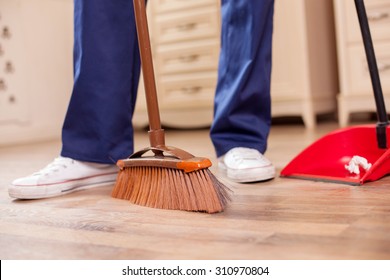 Close Up Of Legs Of Man Standing And Sweeping Flooring. He Is Holding A Broom And Scoop