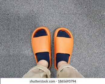 Close Up Legs Of Man Wearing Socks And Orange Rubber Slipper For Indoor Stand On Grey Fabric Floor Background.