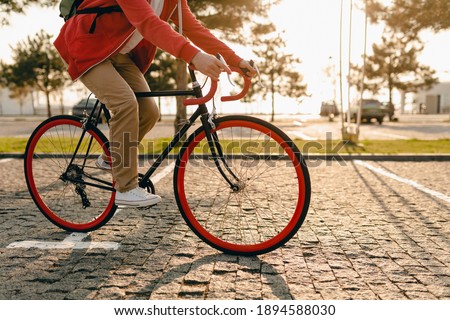 close up legs in sneakers and hands on steering wheel of hipster style bearded man in red hoodie and beige trousers riding alone with backpack on bicycle healthy active lifestyle traveler backpacker