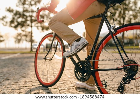 close up legs in sneakers and hands on steering wheel of hipster style bearded man in red hoodie and beige trousers riding alone with backpack on bicycle healthy active lifestyle traveler backpacker