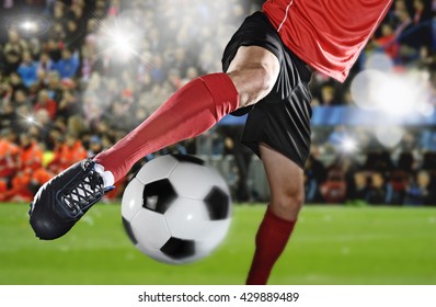 Close Up Legs And Soccer Shoe Of Football Player In Action Kicking Ball Wearing Red Jersey And Sock Playing On Stadium With Audience Flashes  And Lens Flare On The Background 