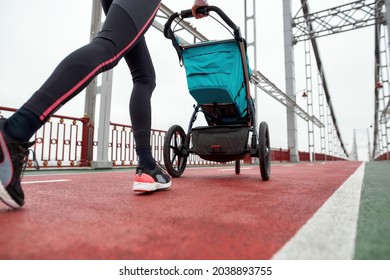 Close Up Of Legs Of Sportswoman Mom Running With A Baby Carriage While Training On A Bridge In The City. Parenting, Healthy Lifestyle, Sports Concept