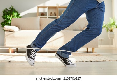 Close Up Of Legs Of Unknown Man Falling Due To What He Stumbles On Power Cord At Home. Man In Jeans And Sneakers Trips Over White Electric Cord While Walking Through Living Room. Low Section.