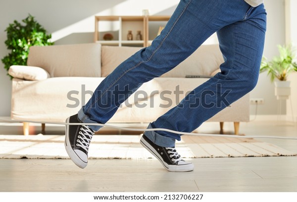 Close up of legs of unknown man falling due to\
what he stumbles on power cord at home. Man in jeans and sneakers\
trips over white electric cord while walking through living room.\
Low section.