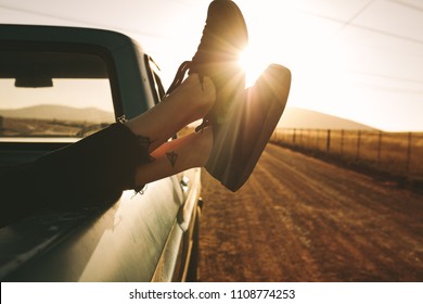 Close Up Of Legs Of A Woman Relaxing At The Back Of A Pick Up Truck On A Highway In Country Side. Legs Hanging Out From Car With Sun In The Background.